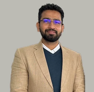Professional headshot portrait of male team member wearing blue glasses and beige business blazer standing against neutral gray background for company about us page